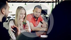 Company of friends sitting at a bar counter in summer day. Beautiful girl taking from table and tasting fresh cocktail