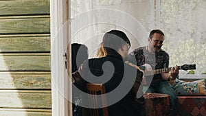 Company of friends at table on terrace of country house. Man play guitar sing