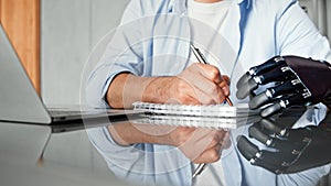 Company manager man with bio hand prothesis makes notes in paper notebook sitting at table