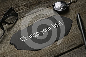Compass,glasses,pen and piece of black paper written with Change Your Life! on wooden background