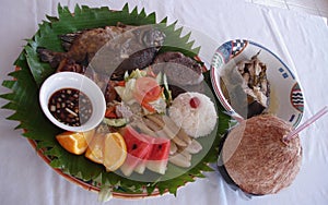 Complete meal of grilled fish, local fruit, rice, coconut juice served on a plate with banana leaf