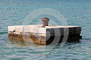 Completely rusted dilapidated iron mooring bollard in middle of stone pier surrounded with calm sea used for tying larger ships