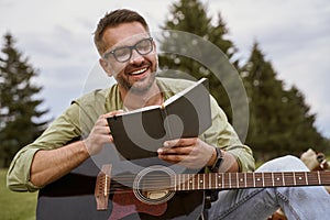 Composing music in nature. Young creative man wearing eyeglasses holding acoustic guitar and writing a song, smiling