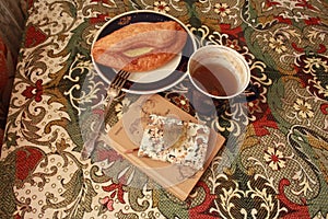 Composition of banana puff on a plate, a blue cup of tea, a fork, two notebooks and a citrine stone on a multi-colored tablecloth.
