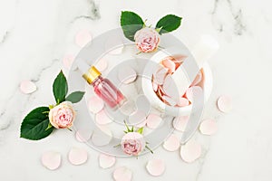 composition with a bottle of rose essential oil, buds and flower leaves, mortar and pestle and rose petals. marble white