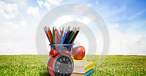 Composition of coloured pencils, books and apple with alarm clock, in sunny field with blue sky