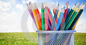 Composition of coloured pencils in mesh metal container, in sunny field with blue sky