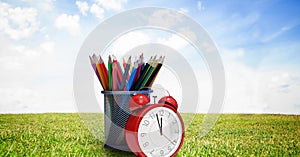 Composition of coloured pencils in pot with alarm clock, in sunny field with blue sky