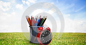Composition of coloured pencils in pot with alarm clock, in sunny field with blue sky