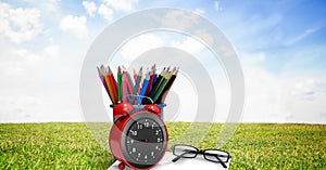 Composition of coloured pencils in pot with glasses and alarm clock, in sunny field with blue sky