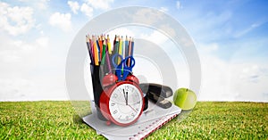 Composition of coloured pencils, sationery and apple with alarm clock, in sunny field with blue sky