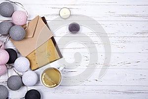 Composition of cup of tea, craft envelope, book and candles, decorated with cotton balls light garland on white wooden background.