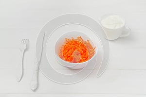 Composition, freshly grated carrot in a plate. On the wooden table, a knife and fork. White background. Light key.