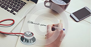 Composition of hand writing thank you note with laptop, coffee, smartphone and stethoscope on desk