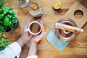 A composition of hands holding cup of tea, teapot and sugar on wooden table. A top view.
