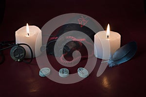 A composition of lighted candles, a bag of runes, an amulet with the image of Yggdrasil and a blue feather on a red background.