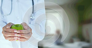 Composition of midsection of female doctor in lab coat with holding apple over out of focus hospital