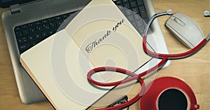 Composition of notebook with thank you note with laptop, coffee cup and stethoscope on wooden desk