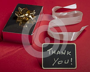 Composition on a red background of a gift with cockade, ribbon and blackboard with the words `thank you`