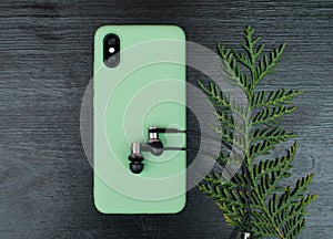 Composition with smartphone in unbranded mint / teal color case, headphones, green leaves of white cedar branch on black wooden ta