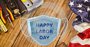 Composition of text happy labor day on face mask with tools and american flag on wood background