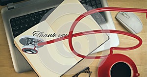 Composition of thank you text on notebook over laptop and heart shaped stethoscope