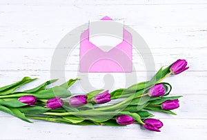 Composition of white blank card in purple color opened craft envelope decorated with purple tulips on white wooden background.