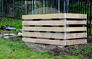 A composter made of natural beech prisms built horizontally with black foil inside in a meadow near a dark wooden fence.