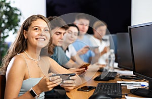 At computer class, young girl student sits with mobile phone and listens admiringly to teacher