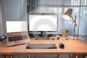 Computer device with blank monitor, laptop, table lamp, keyboard, mouse on wooden desk in home office