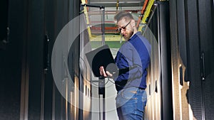 Computer engineer checks computers in server room, using laptop.