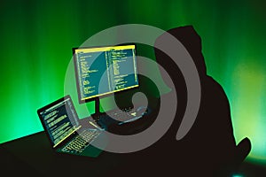 Computer hacker silhouette of hooded man typing on computer in a dark green room.