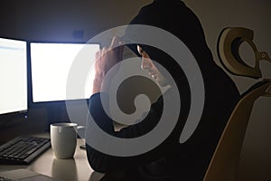 Computer hacker. Young man in black hoodie using multiple computers for stealing data while sitting in dark room