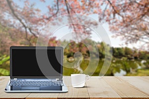 Computer laptop with black screen and hot coffee cup on wooden table top on blurred lake and cherry blossom tree background