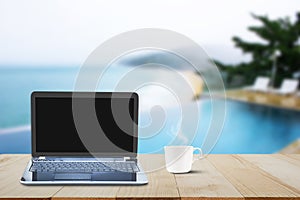 Computer laptop with black screen and hot coffee cup on wooden table top on blurred pool and beach background