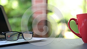 Computer laptop with reading glasses and coffee in red cup with garden background. Work from home