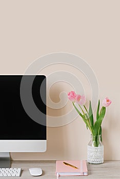 A computer monitor, a bouquet of pink tulips, and a notebook with a pen on a light wooden table against a beige wall. The
