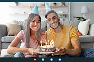 Computer screen view of happy multiracial couple showing birthday cake with candles at webcam, celebrating b-day online