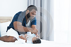 Concentrated African Nigerian bearded father changing diaper and clothes for newborn baby lying on white bed in bedroom. Single