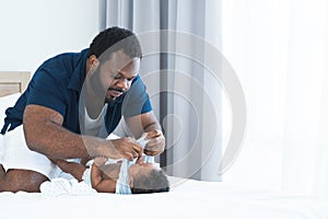 Concentrated African Nigerian bearded father changing diaper and clothes for newborn baby lying on white bed in bedroom. Single