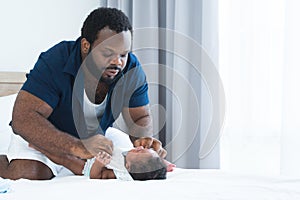 Concentrated African Nigerian bearded father changing diaper and clothes for newborn baby lying on white bed in bedroom. Single