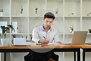 Concentrated asian businessman sitting front of laptop and writing information on notebook
