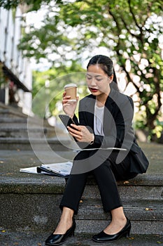 A concentrated Asian businesswoman is responding to messages on her phone on the stairs outdoors