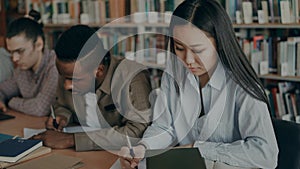 Concentrated asian girl student preparing for examination and writing notes while sitting at table at university library