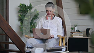 Concentrated Asian man typing on laptop keyboard sitting in living room indoors. Focused young handsome Chinese guy