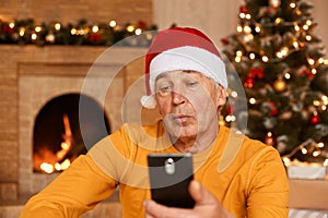 Concentrated attentive mature man wearing orange sweater and santa claus hat using mobile phone while posing at home in decorated