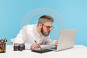 Concentrated bearded man designer drawing on professional tablet looking at laptop display, creating project sitting at workplace