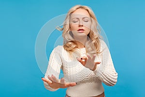 Concentrated blond woman in white knitted sweater trying to move with closed eyes and raised hands, vision problems, blindness