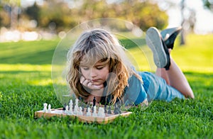 Concentrated boy developing chess strategy, playing chess board game. Cute little boy playing chess, laying on grass in