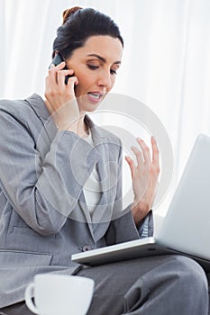 Concentrated businesswoman calling with her mobile phone and using laptop sitting on sofa